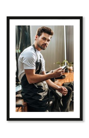 handsome young hairstylist using smartphone and looking at camera in beauty salon