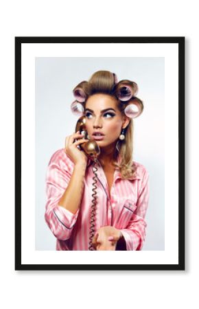A beautiful young showy girl with bright pin-up make-up speaks on a golden dial telephone in pink striped silk pajamas and pink curlers.
