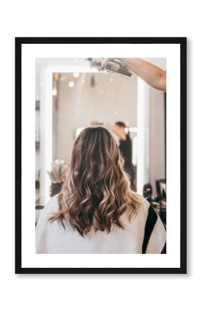 Beautiful young woman with long curly hair in hair salon. Professional hairdresser styling with hairspray.