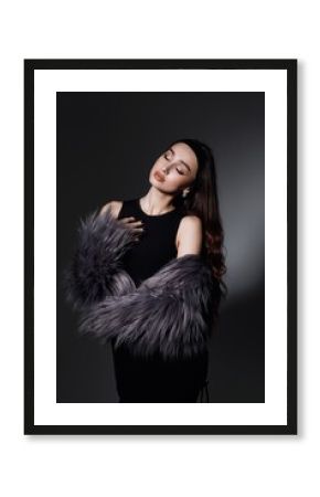 girl in the studio with long brown hair falling freely on her shoulders. She is dressed in a black top and has a fluffy gray fur coat draped over her shoulders.