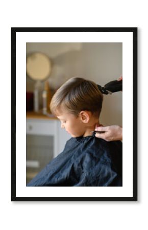 A little blond boy gets a haircut with a hair clipper, sitting in a salon chair with a cape. The hairdresser carefully trims his hair. The child is calm and enjoying the experience. 