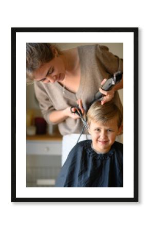 A little blond boy gets a haircut, sitting in a salon chair with a cape. The hairdresser holds scissors, a comb, and a hair clipper. The child is calm and enjoying the experience. 