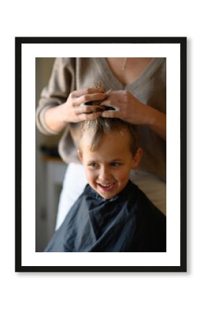 A little blond boy enjoys his new haircut as the hairdresser styles his hair with mousse. He is happy, laughing, and having fun in the salon chair. Concept of childhood grooming and self-care.
