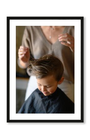A little blond boy enjoys his new haircut as the hairdresser styles his hair with mousse. He is happy, laughing, and having fun in the salon chair. Concept of childhood grooming and self-care.