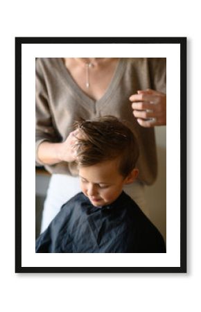 A little blond boy enjoys his new haircut as the hairdresser styles his hair with mousse. He is happy, laughing, and having fun in the salon chair. Concept of childhood grooming and self-care.