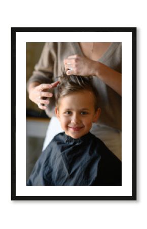 A little blond boy enjoys his new haircut as the hairdresser styles his hair with mousse. He is happy, laughing, and having fun in the salon chair. Concept of childhood grooming and self-care.