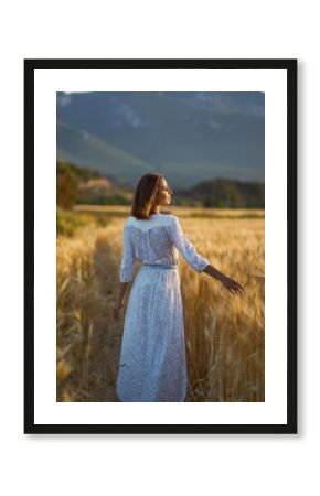Beautiful girl in long white dress on the wheat field enjoying golden sunset outdoors. Her hand touching of spikes