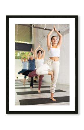 Positive European young woman standing in Vrksasana Tree pose on mats during group yoga class in gym