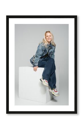 full length of cheerful blonde woman in blue denim outfit and trendy sneakers sitting on white cube on grey.