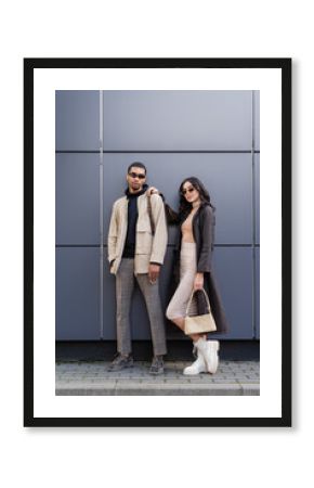 full length of stylish interracial couple in autumnal outfits and trendy sunglasses standing near building.