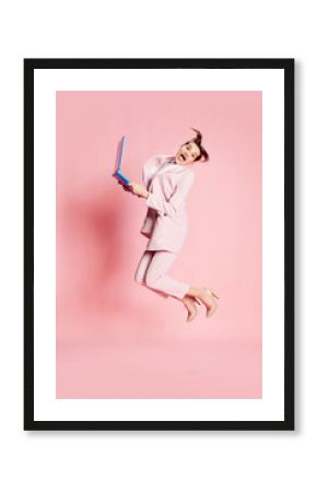 Portrait of beautiful young girl in suit and glasses jumping with laptop over pink background. Professional energy