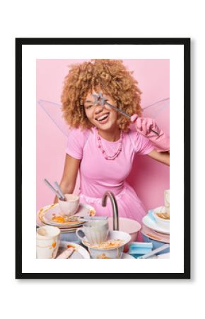 Vertical shot of cheerful curly haired female fairy covers eye with magic wand smiles happily going to wash up maintains housekeeping has good mood isolated over pink background. Domestic chores