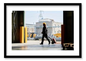 Worker moving loaded hand truck in industrial warehouse during daytime with visible heavy machinery and industrial equipment in the background
