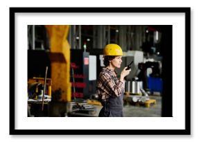 Young engineer wearing hard hat, holding radio device while standing in industrial workshop