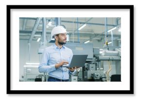 Chief Engineer in the Hard Hat Walks Through Light Modern Factory While Holding Laptop. Successful, Handsome Man in Modern Industrial Environment.