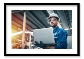 Confident engineer in blue jumpsuit holding laptop computer in a warehouse.