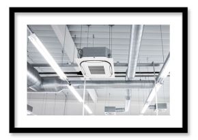 Ceiling mounted cassette type air condition units with other parts of ventilation system (tubes, cables and vents) located inside commercial hall with hanging lights and other construction parts.