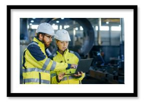 Male and Female Industrial Engineers in Hard Hats Discuss New Project while Using Laptop. They Make Showing Gestures.They Work in a Heavy Industry Manufacturing Factory.