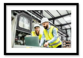 A portrait of an industrial man and woman engineer with laptop in a factory, working.