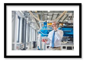 Male supervisor digital tablet talking on cell phone next to plastic industrial molder machinery in factory