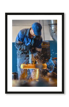 Male worker grinding metal pipe in industrial setting with safety gear.