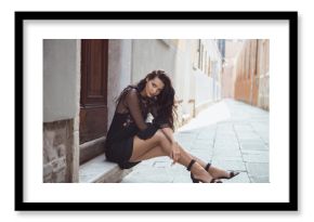 Travel tourist girl on vacation posing on city street with beautiful legs in high heel shoes. Attractive young romantic passion woman sitting on sidewalk against beautiful view on venetian quiet