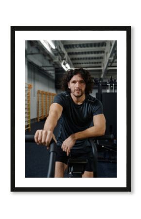 Vertical medium long portrait of confident Caucasian man posing for camera while using cycle bike in modern gym