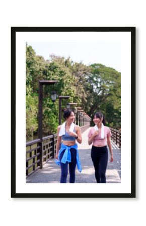 Two young Asian women walking and chatting on a wooden bridge after exercising together in a scenic park