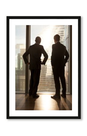 Vertical rear view of afro american and caucasian businessmen in formalwear stand near large window, look at big city buildings outside, two diverse multi-ethnic partners silhouettes deep in thought