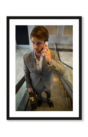 Business professional climbing escalator with railings in lobby, holding smartphone and briefcase