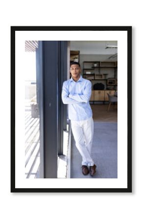 Confident businessman in blue shirt standing by office window, looking thoughtful