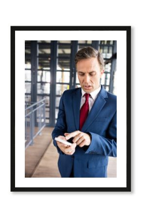 Businessman standing in office corridor using smartphone near glass partitions on wood floor