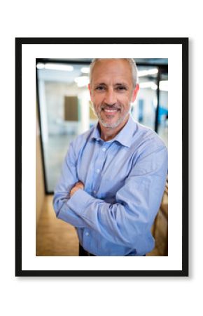 Senior businessman standing in modern office hallway, smiling with arms crossed by glass partitions