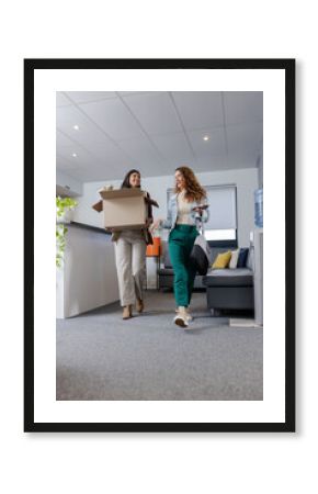 Two coworkers walking in office lounge carrying box and holding smartphone and bag near counter