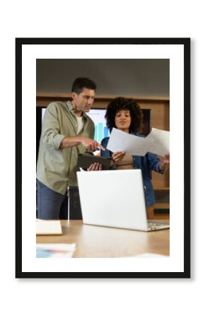 Diverse colleagues pointing at tablet and reviewing printed charts in modern office, with laptop