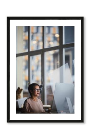 Young modern businesswoman sitting by table in office and browsing in the net