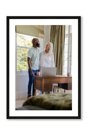 Standing Diverse senior couple sharing coffee behind desk in office with laptop and ceramic mugs