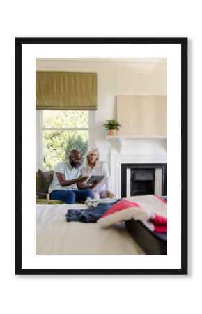 Sitting diverse couple reviewing trip plans on tablet on green sofa in bedroom, with open suitcase