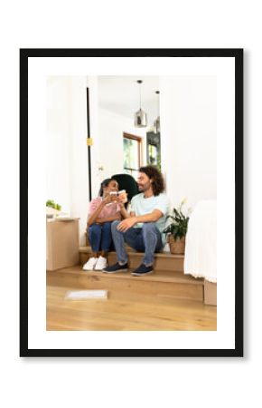 Young diverse couple sitting on stairs, enjoying coffee break while unpacking boxes at home
