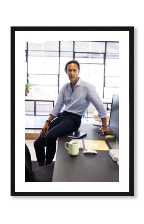 Confident businessman sitting on desk in modern office, looking at camera
