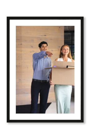 Colleagues smiling and pointing while carrying box in modern office hallway