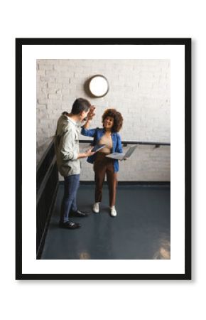 High-fiving diverse colleagues holding laptop and tablet on office stair landing, with wall light