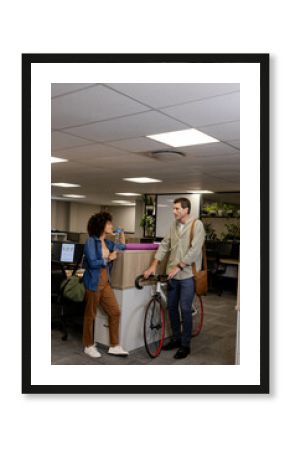 Chatting diverse coworkers leaning against desk in open-plan office, with bicycle and water bottle