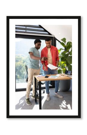 Reviewing printed charts Diverse coworkers standing at modern office desk, with laptop and papers