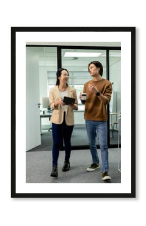 Asian coworkers wearing sweater and blazer walking in office holding tablet and reusable coffee cup