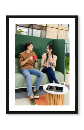 Asian colleagues chatting over coffee while sitting on green booth at office lounge with tablet