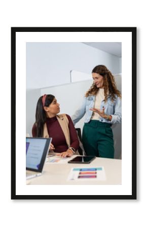 Diverse female colleagues discussing project on laptop, tablet and charts at desk in office cubicle