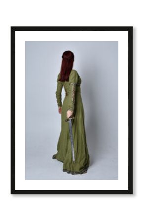 full length portrait of red haired girl wearing celtic, green medieval gown. Standing pose, holding a sword, isolated against a studio background.