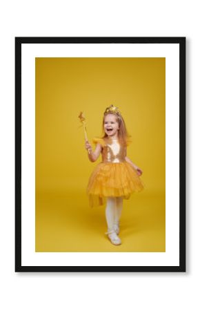 Joyful little girl with long hair in a tulle golden dress and princess crown holding a magic wand  on yellow background. Celebrating a colorful carnival for kids, expressing positive birthday