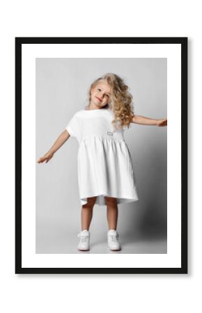 Little blonde curly positive princess girl in white casual dress and sneakers standing dancing walking and smiling  over grey wall